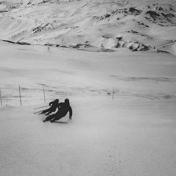 Schwarz-Weiß-Foto: Zwei Skifahrer carven synchron einen weiten Hang hinab. Im Hintergrund raue, schneebedeckte Berge.